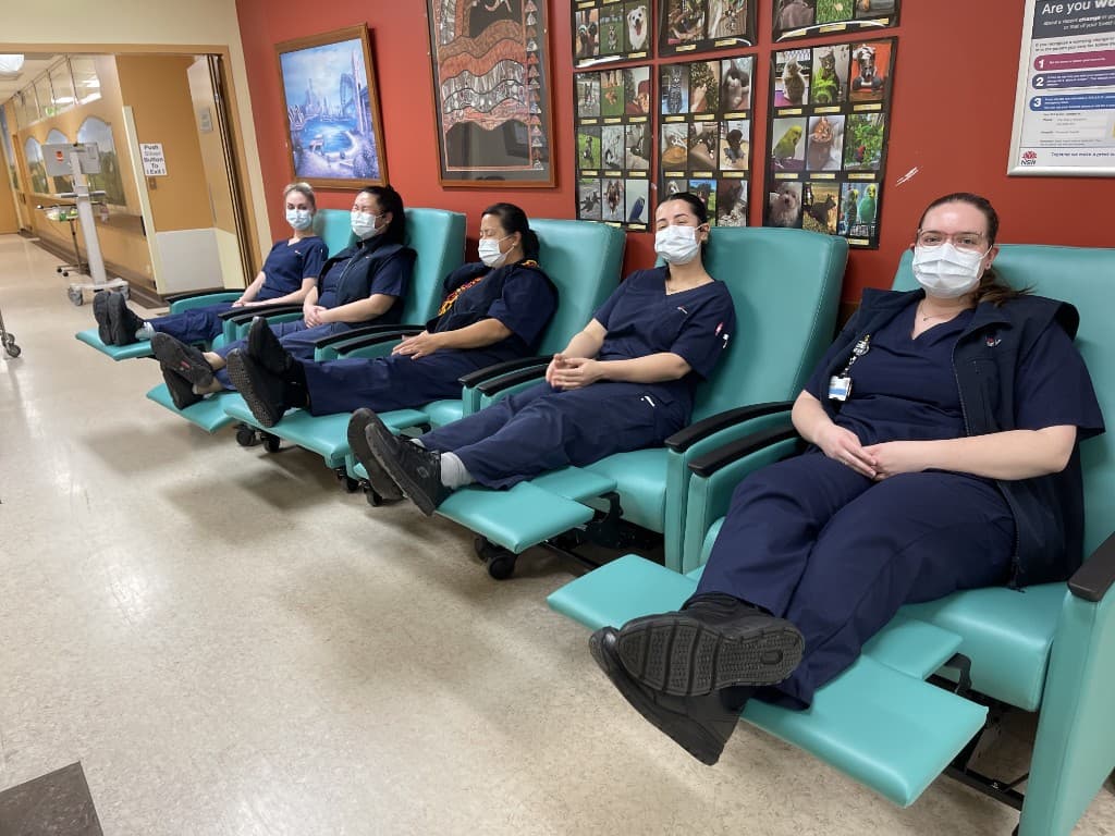 Five Ward C5A nurses sitting in the new teal recliner chairs in a hospital corridor, putting them through quality-control testing before the chairs go into patient rooms